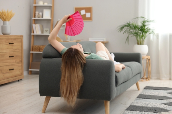 Woman using pink hand fan to cool herself while lying down on the couch at home